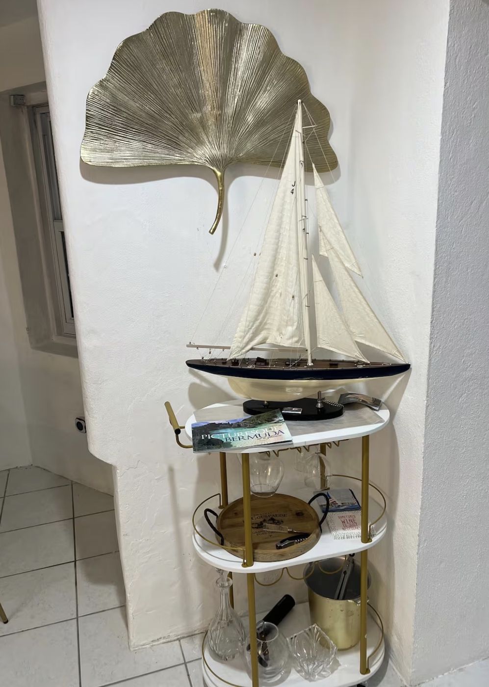 Gold and white bar cart beneath a model sailboat and gold ginkgo leaf