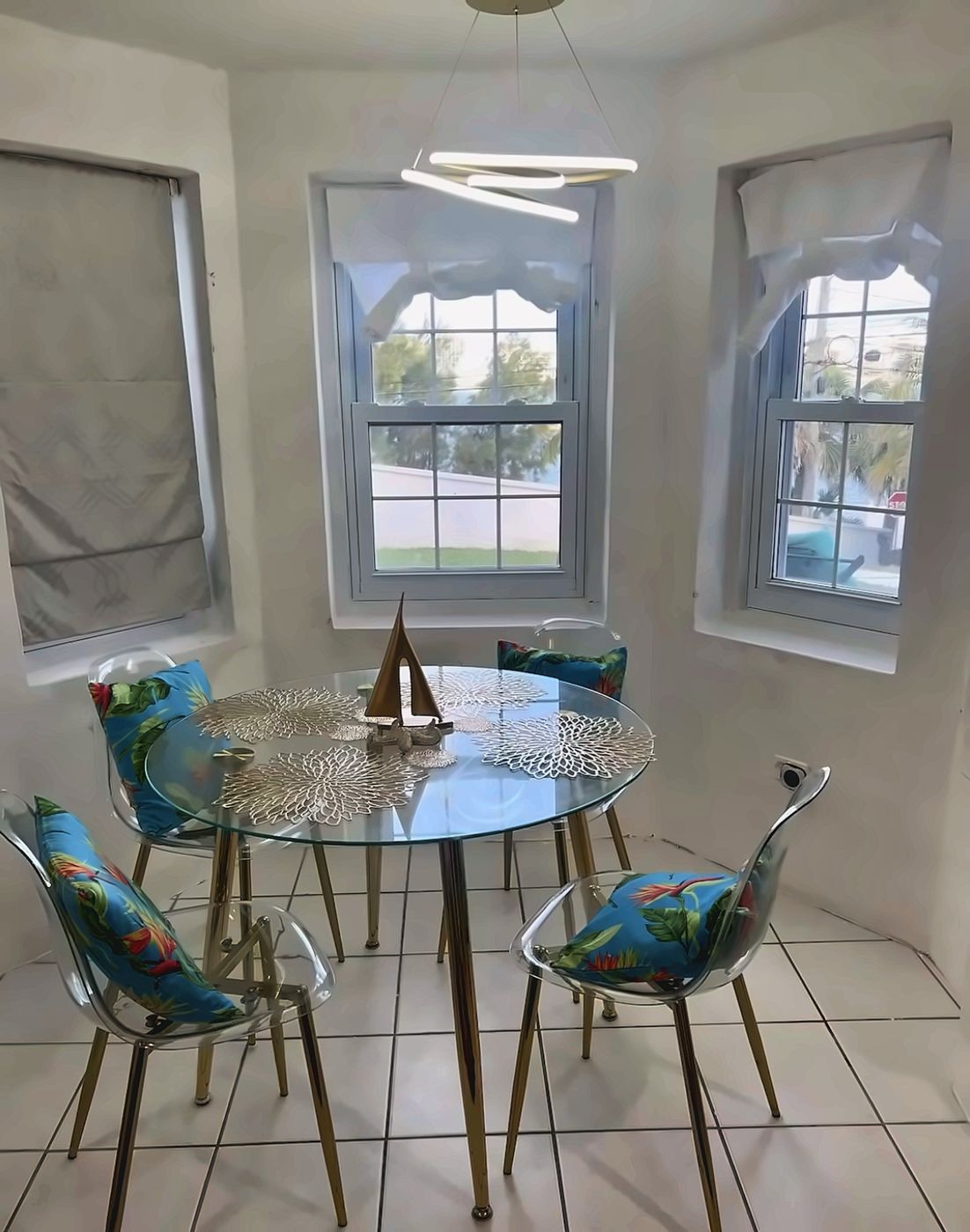 Round glass dining table with four clear chairs and tropical cushions in a bay-window nook